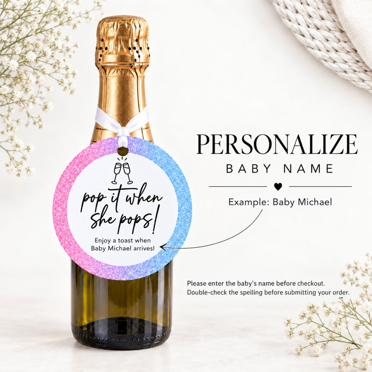 A personalized bottle tag sticker shown attached to a champagne bottle, with text reading 'PERSONALIZE BABY NAME' and 'pop it when she pops!'. The circular tag has a pink-to-blue glitter gradient and is displayed on a light background with decorative flowers and a woven textile.
