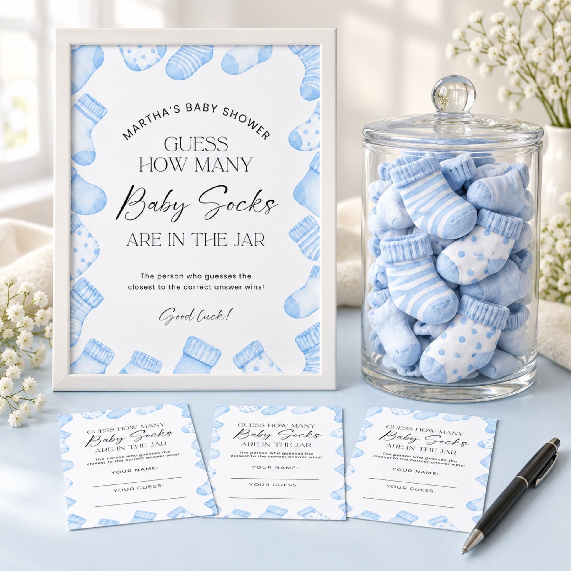 Baby shower game set featuring a white framed sign with blue baby sock illustrations, a glass jar filled with blue and white striped baby socks, and three matching participation cards on a light surface with floral props.
