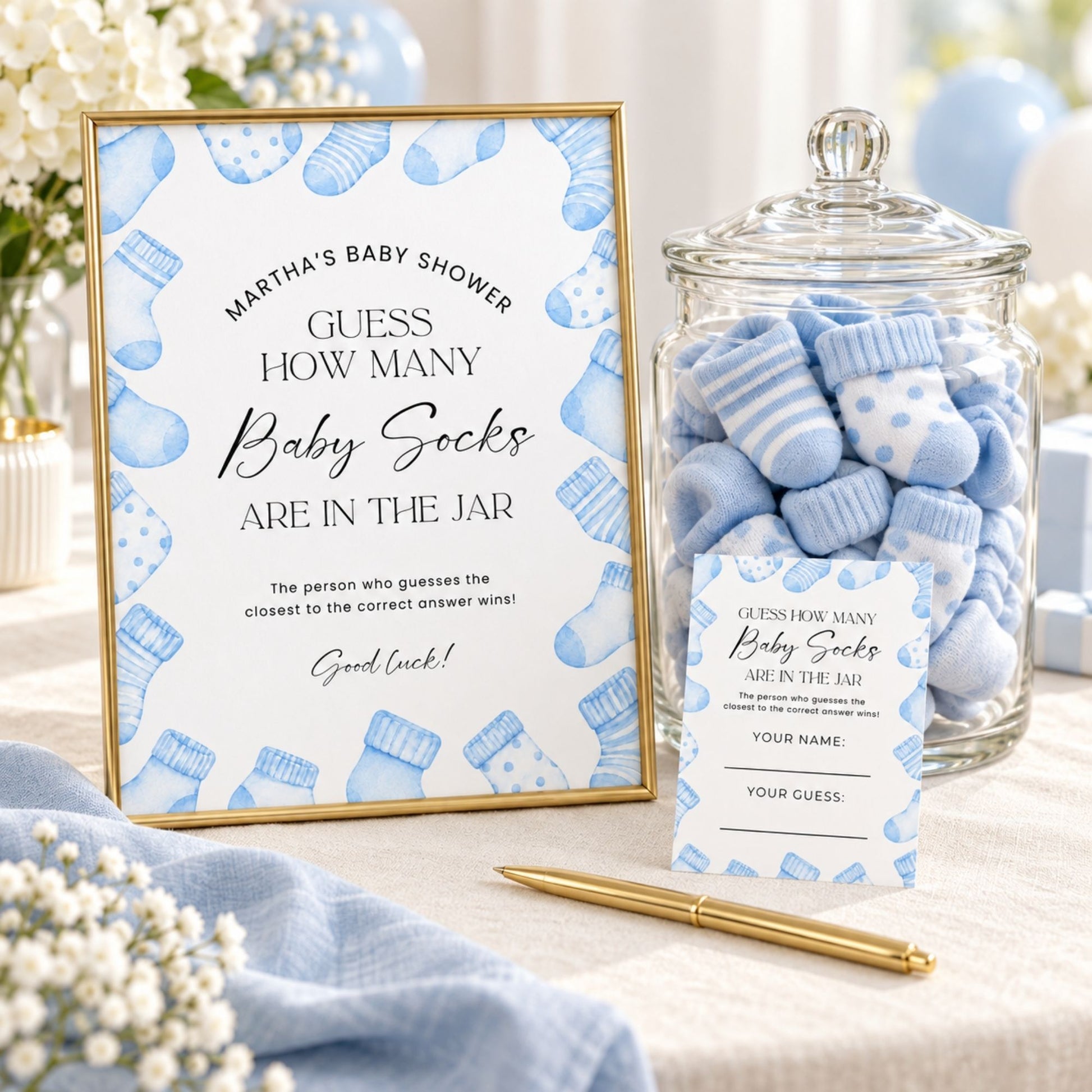 Baby shower game setup with a framed sign reading 'Guess how many baby socks are in the jar,' a glass jar filled with blue-and-white baby socks, a gold pen, and a card in a softly styled indoor setting.