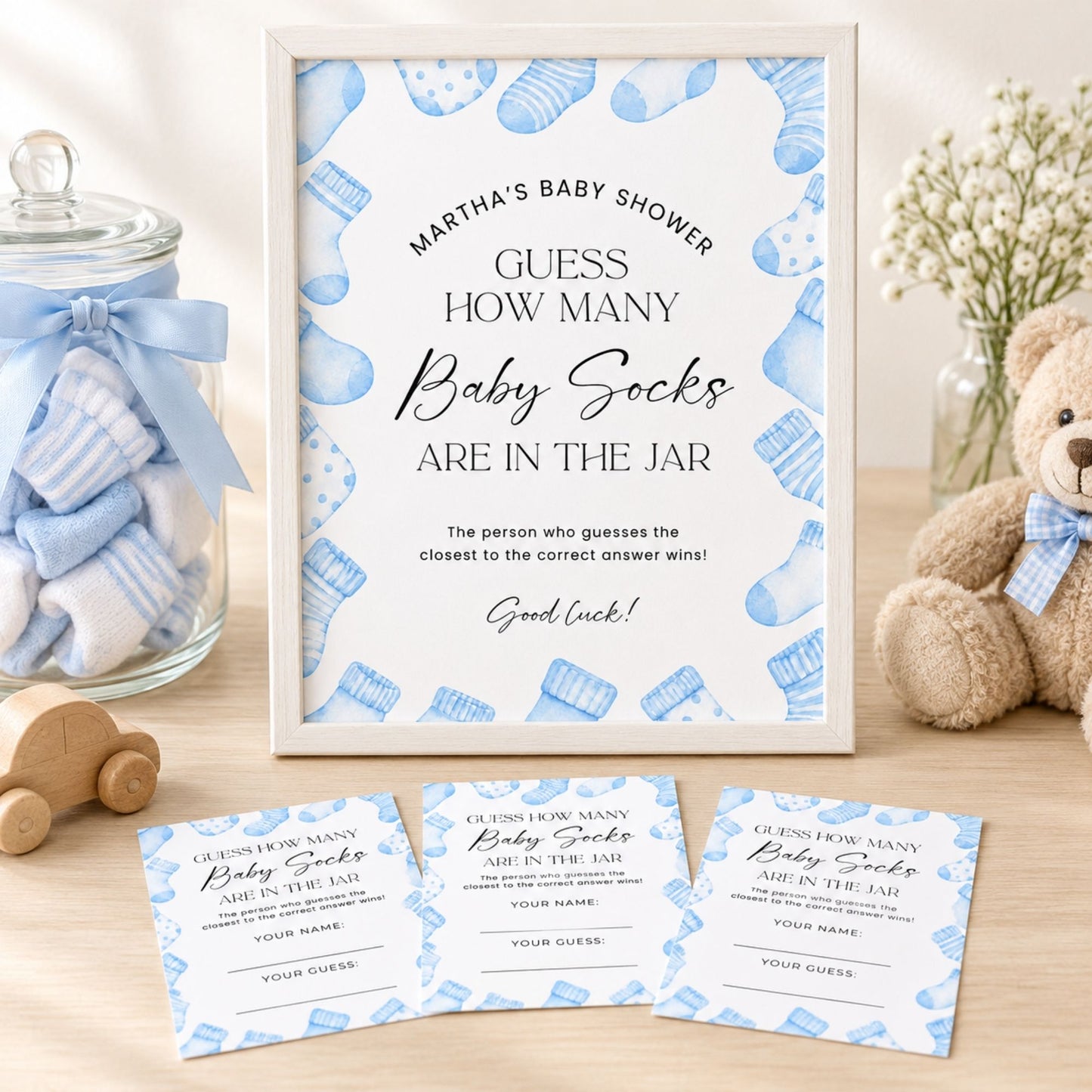 A framed baby shower game card reading 'MARTHA'S BABY SHOWER GUESS HOW MANY Baby Socks ARE IN THE JAR' is displayed on a tabletop, with three matching cards in front and blue-and-white baby socks visible inside a glass jar behind it. A teddy bear, flowers, and a wooden toy are also present in a styled indoor setting.