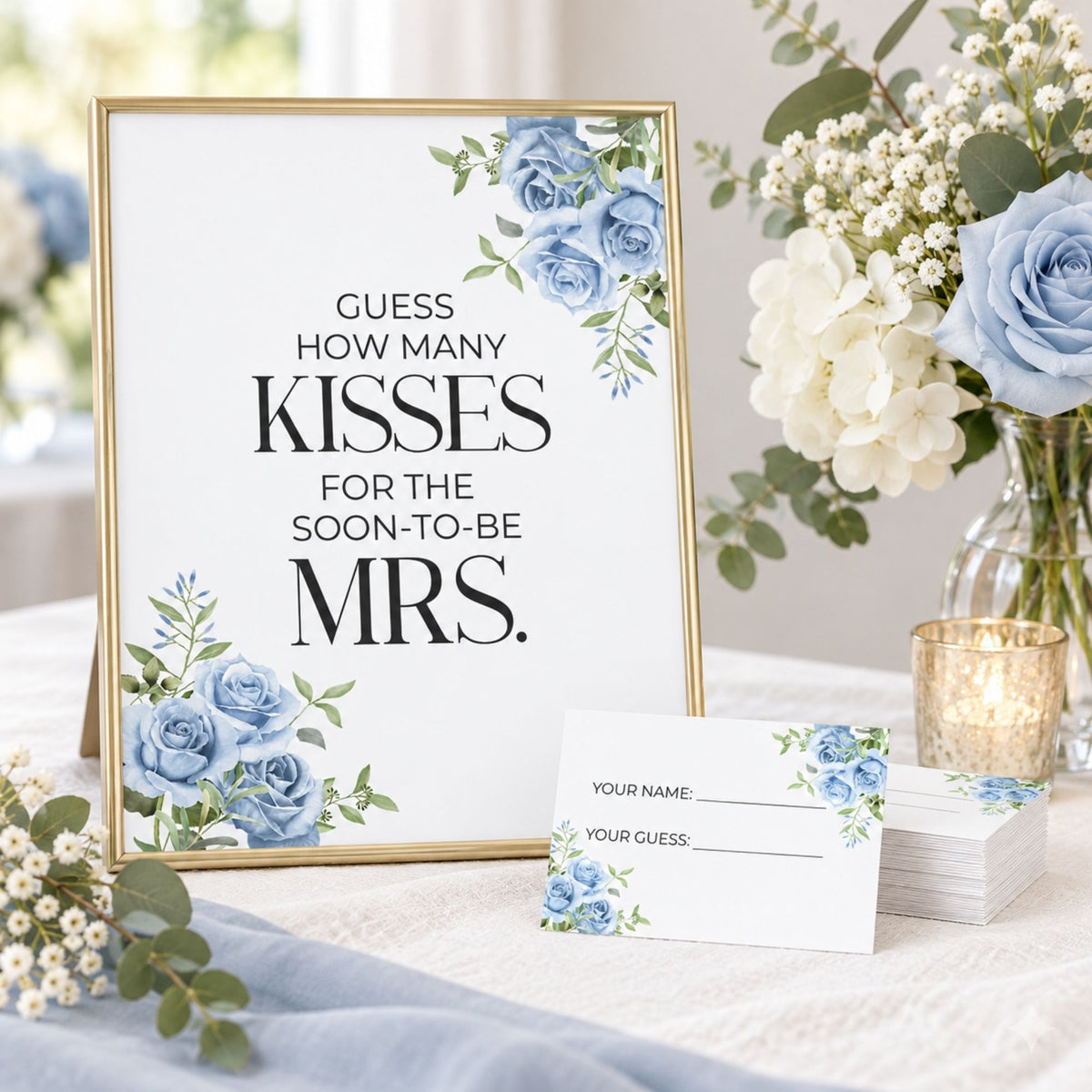 A framed wedding sign with blue floral corner accents and the text 'GUESS HOW MANY KISSES FOR THE SOON-TO-BE MRS.' stands on a table beside matching printed cards and a glass candle holder, with flowers in the background.