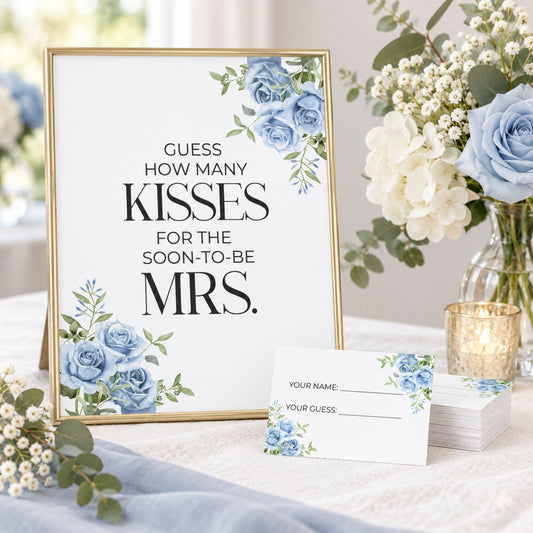 A framed wedding sign with blue floral corner accents and the text 'GUESS HOW MANY KISSES FOR THE SOON-TO-BE MRS.' stands on a table beside matching printed cards and a glass candle holder, with flowers in the background.