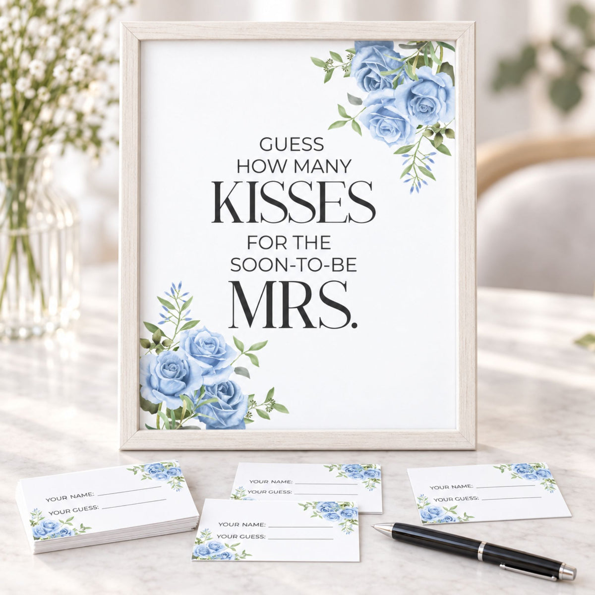 A framed wedding reception sign reading 'GUESS HOW MANY KISSES FOR THE SOON-TO-BE MRS.' with blue rose illustrations, displayed on a marble table with matching name cards and a pen in front.