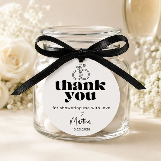 A clear glass jar candle tied with a black ribbon and a round white tag that reads 'thank you for showering me with love' and is personalized with the name 'Martha' and a date. The jar is filled with white candy and is styled with white roses in a warm, soft-focus setting.
