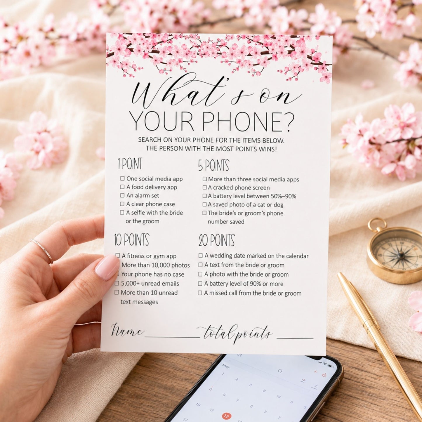 A hand holds a printed checklist card titled "What's on YOUR PHONE?" with pink cherry blossom graphics at the top, photographed over a wooden surface with blurred pink blossoms, a gold compass, and a smartphone in the background.