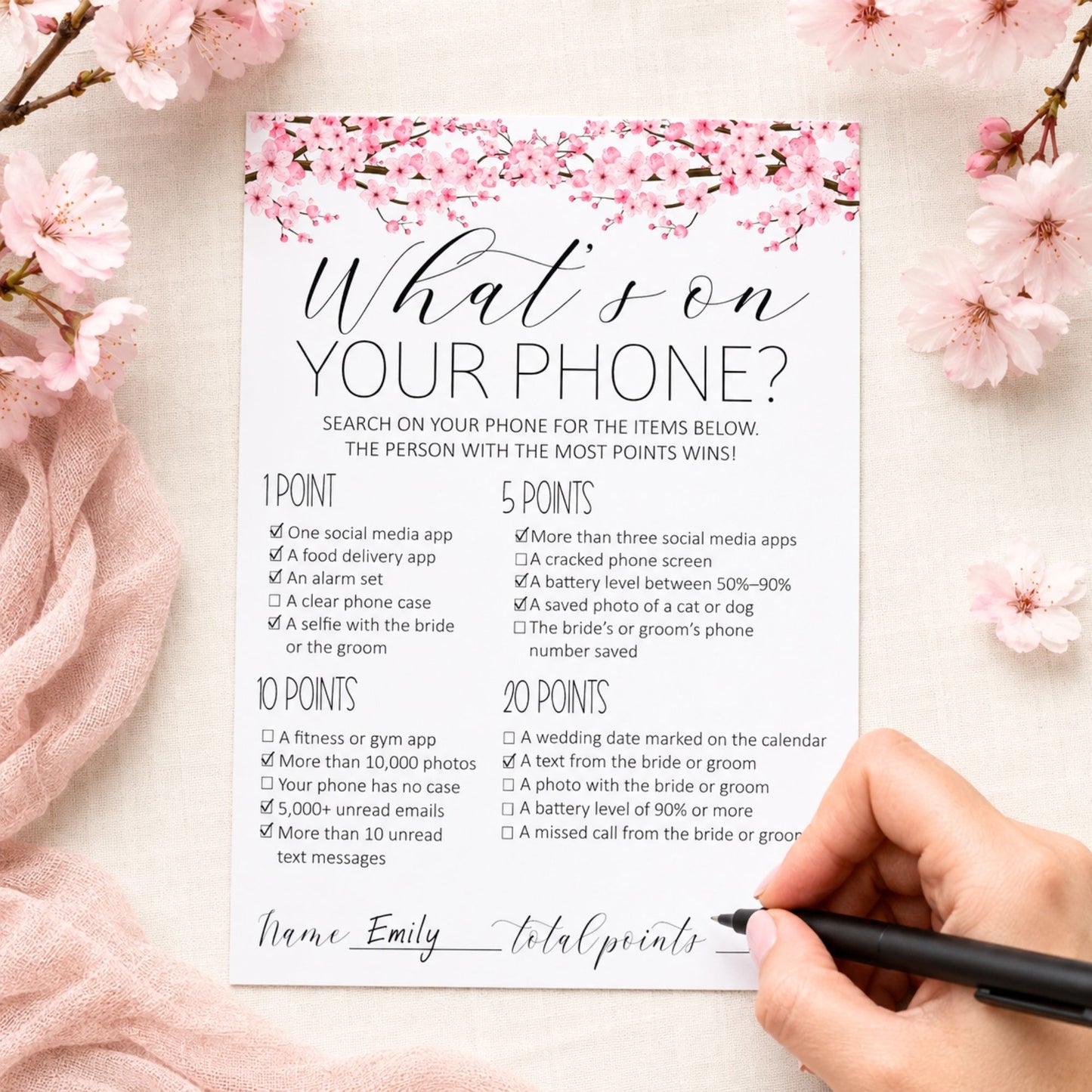 A printed phone-related checklist card with pink cherry blossom graphics at the top, shown on a pink fabric background with cherry blossom branches around the edges and a hand holding a black pen at the bottom right.