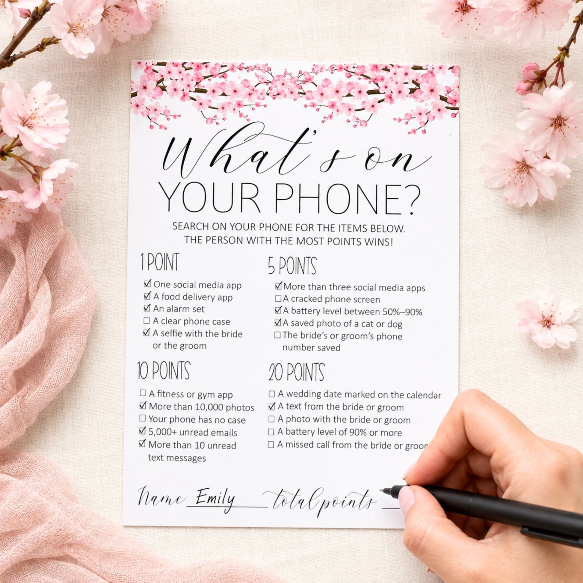 A printed phone-related checklist card with pink cherry blossom graphics at the top, shown on a pink fabric background with cherry blossom branches around the edges and a hand holding a black pen at the bottom right.
