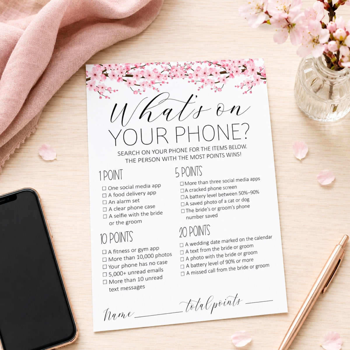 A printed checklist card titled "What's on YOUR PHONE?" styled on a light wood surface with pink blossoms, a pink fabric cloth, a smartphone, and a rose gold pen. The card contains numbered points and a name line for total points.