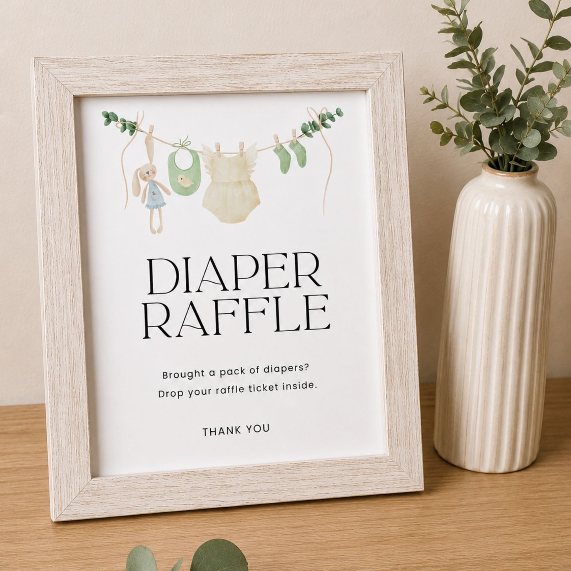 Framed 'Diaper Raffle' poster with illustrations of baby items on a string, placed on a wooden surface next to a vase.