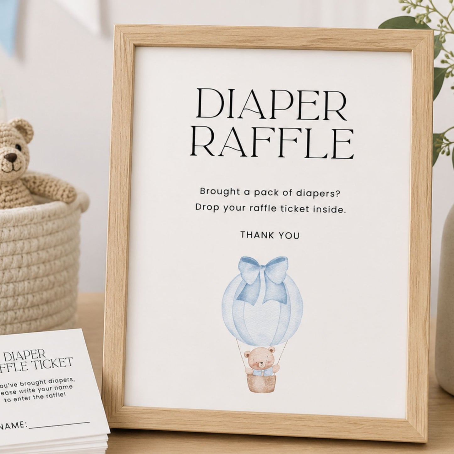 Framed baby shower raffle sign with teddy bear and balloon illustration, next to a basket and card.