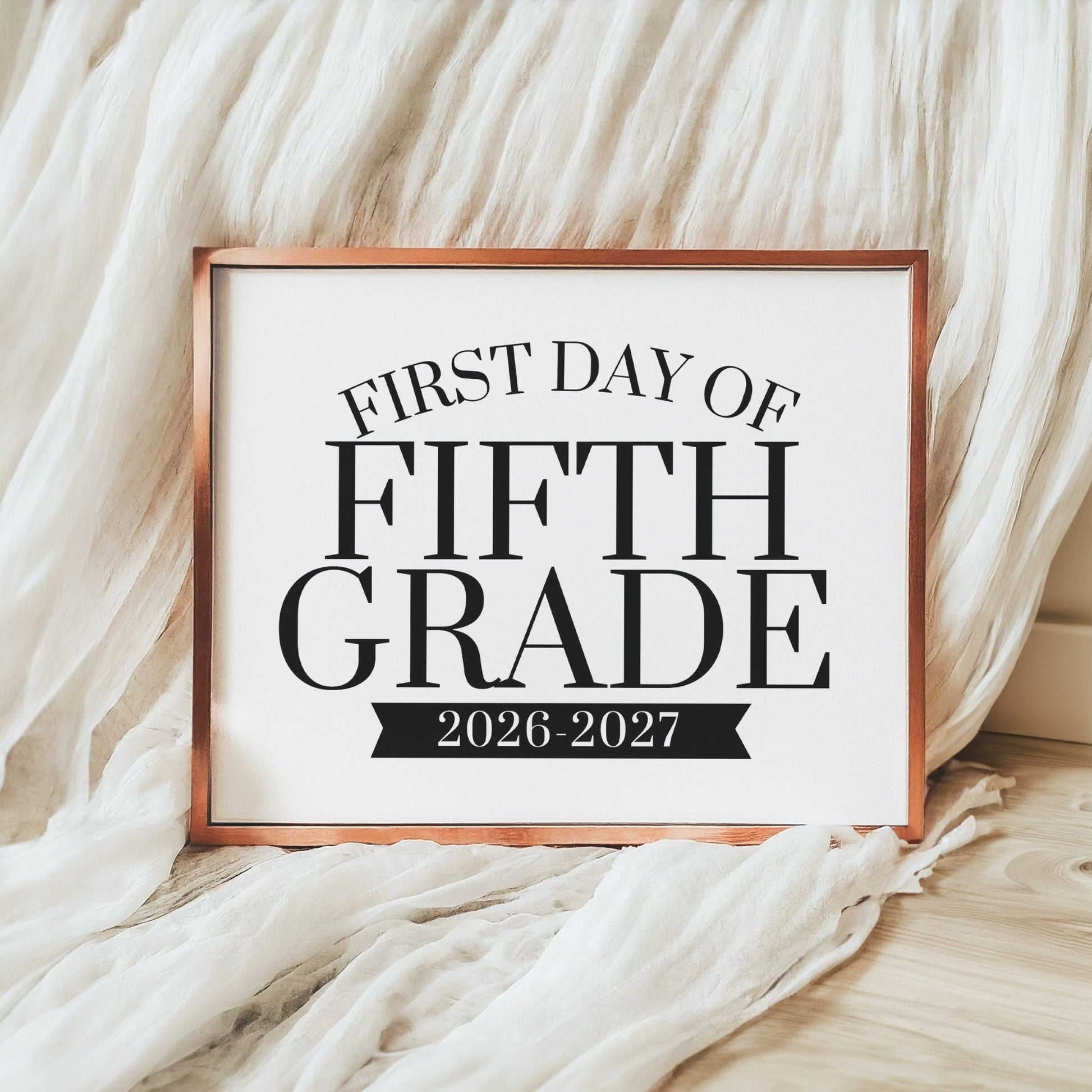 A framed typographic sign reading 'FIRST DAY OF FIFTH GRADE 2026-2027' in black text on a white background, with a copper-toned frame, styled against draped white fabric and a light wood floor.