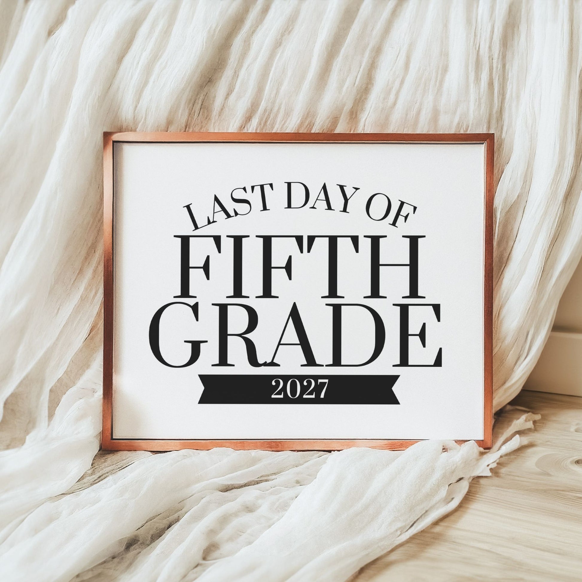 A framed typographic sign reading 'FIRST DAY OF FIFTH GRADE 2026-2027' in black text on a white background, with a copper-toned frame, styled against draped white fabric and a light wood floor.
