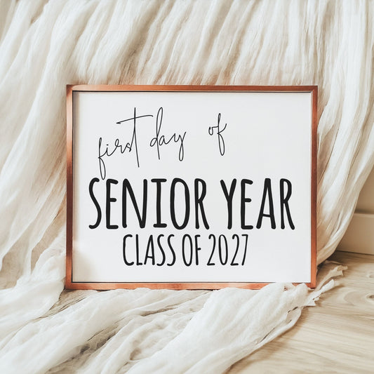 A framed sign reading 'first day of SENIOR YEAR CLASS OF 2027' in black lettering on a white background, with a copper-toned frame, styled on a light wood floor against draped white fabric.