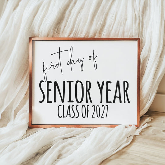 A square framed sign with a copper-toned frame and white background reading 'first day of SENIOR YEAR CLASS OF 2027' in black lettering, styled on a light wood floor with draped white fabric.