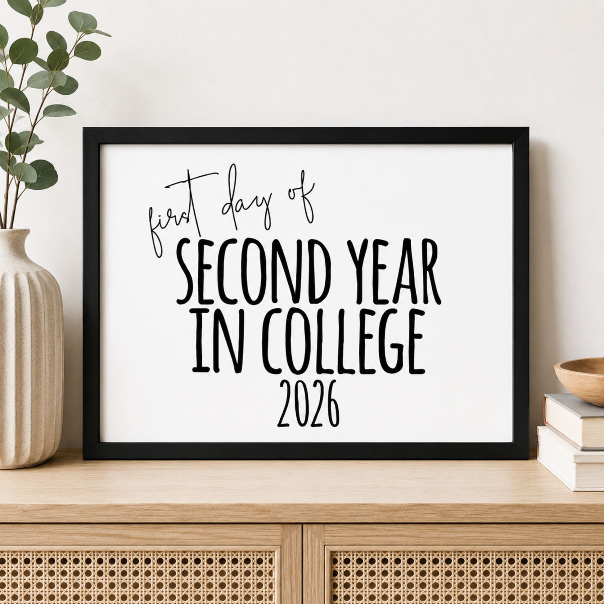 A framed typographic wall print reading 'first day of SECOND YEAR IN COLLEGE 2026' in black text on a white background, displayed on a light wood cabinet with a vase and books nearby.