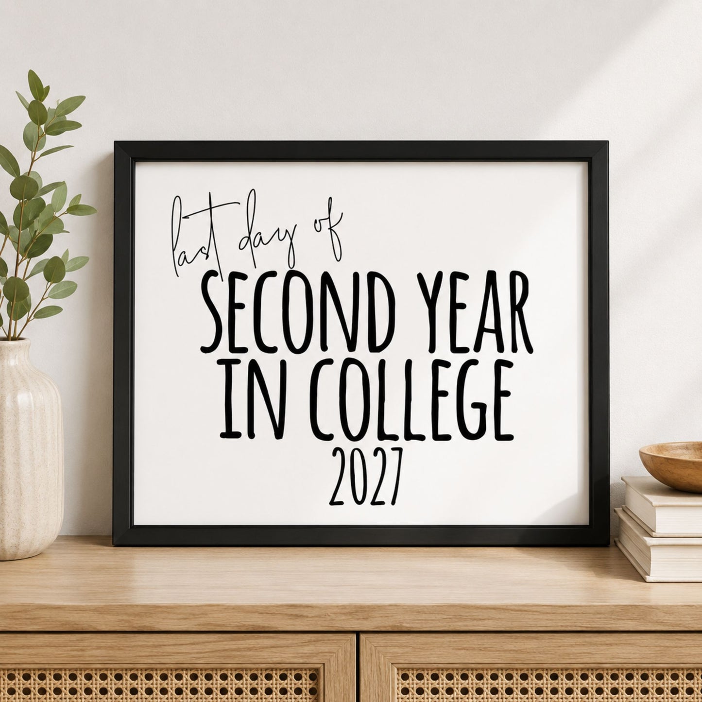 A framed typographic print reading 'last day of SECOND YEAR IN COLLEGE 2027' in black lettering on a white background, displayed on a light wood cabinet with a vase and books nearby.