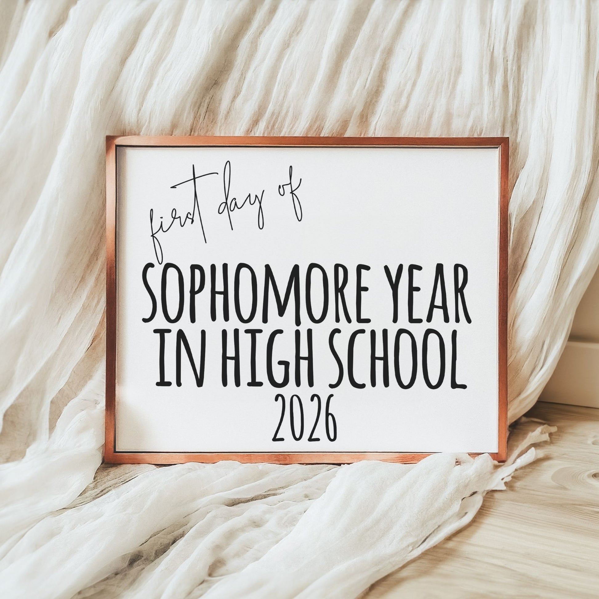 A square framed sign with a copper-toned frame and white background reading 'first day of SOPHOMORE YEAR IN HIGH SCHOOL 2026' in black lettering, styled on a light wood floor with draped white fabric.