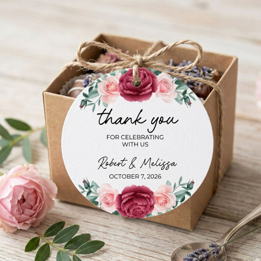 A square kraft paper gift box tied with a twine bow, featuring a round white 'thank you' label with pink and burgundy floral artwork and personalized wedding text. The box is styled on a light wood surface with pink flowers, green leaves, and a spoon with lavender.