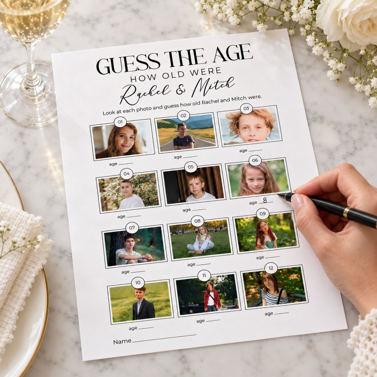 A printed 'Guess the Age' photo puzzle sheet on a marble surface, surrounded by flowers and a wine glass. A hand holds a pen next to the sheet, which features photos of children labeled with ages and the names Rachel & Mitch.