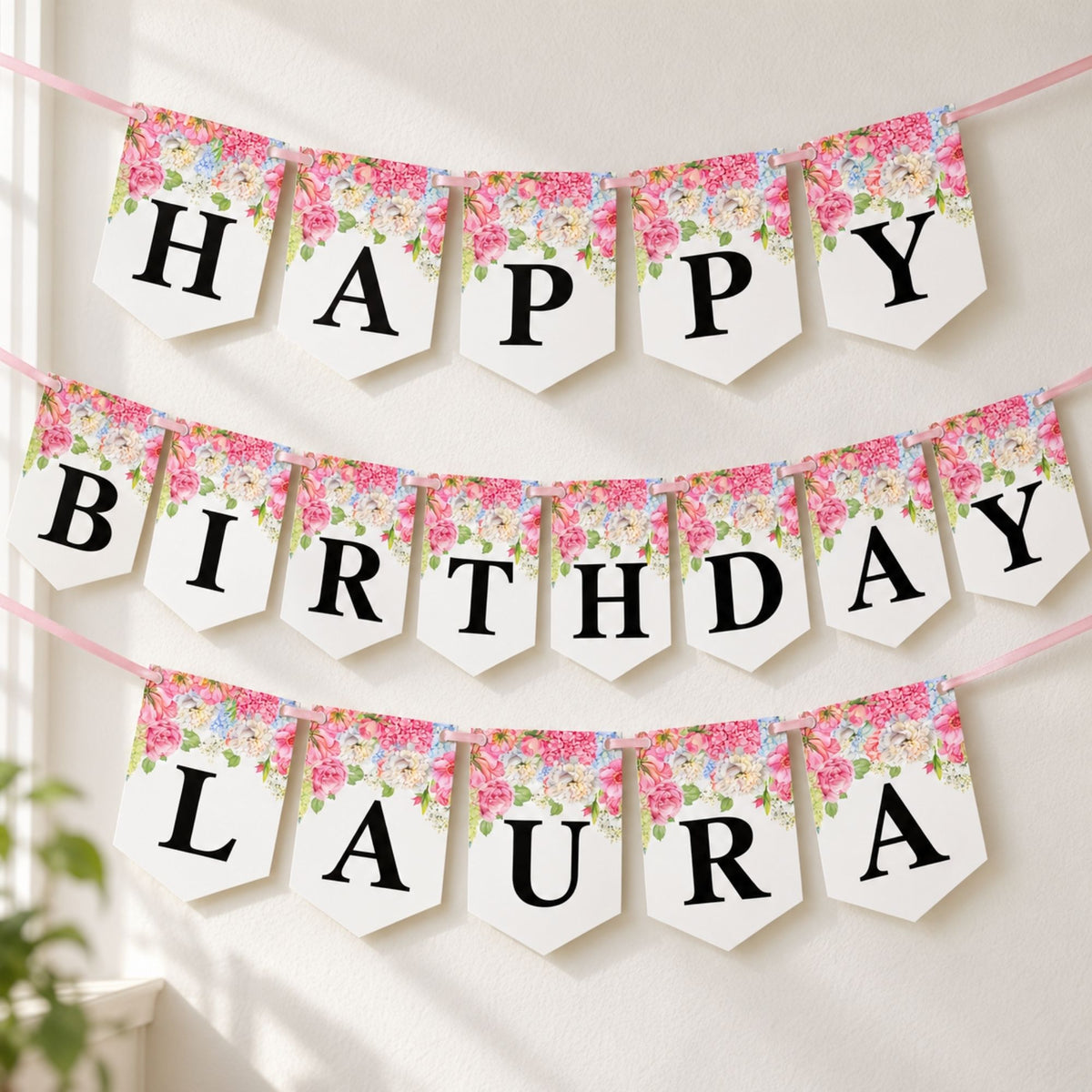 Personalized 'HAPPY BIRTHDAY LAURA' party banner with white hexagonal flags, black lettering, and a pink floral print, hanging on a wall.