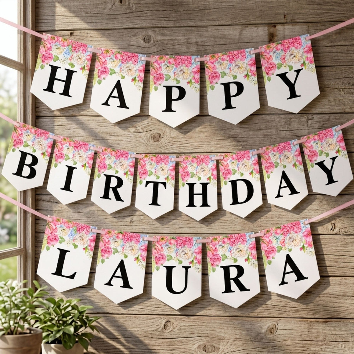 Personalized 'HAPPY BIRTHDAY LAURA' banner with floral pennants hanging on a rustic wooden wall, with a window and potted plants visible on the left.