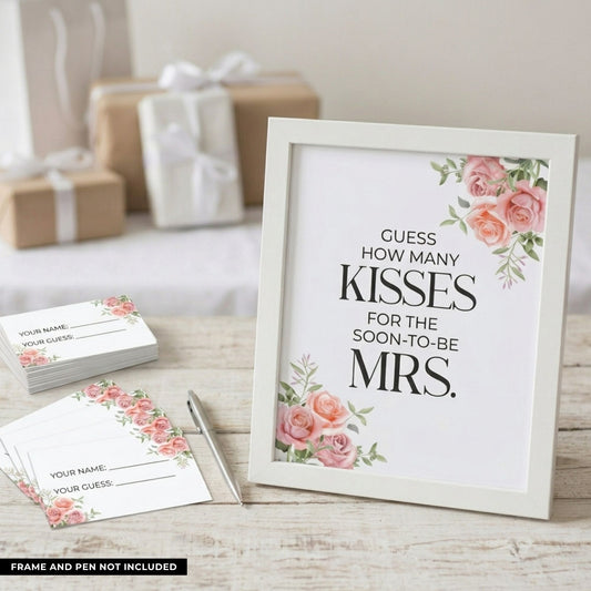 A white framed wedding sign with pink rose illustrations and the text 'GUESS HOW MANY KISSES FOR THE SOON-TO-BE MRS.' displayed on a light wood surface, with matching printed cards and a pen nearby and gift boxes blurred in the background.
