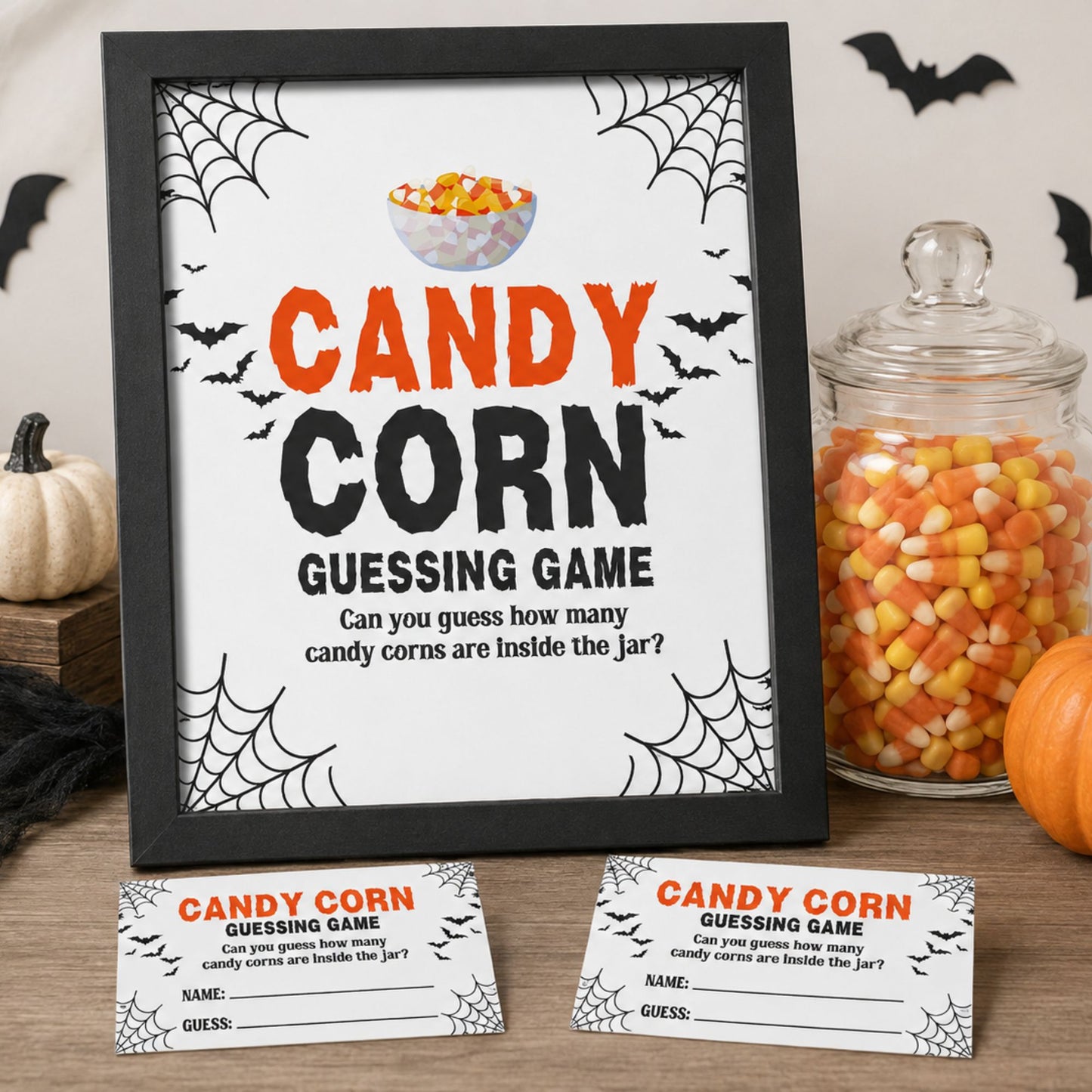 A framed Halloween-themed 'Candy Corn Guessing Game' sign displayed on a tabletop, with two matching game cards in front and a glass jar of candy corn beside it. The sign has spiderwebs, bats, and a bowl of candy corn illustration on a white background, with orange and black text.