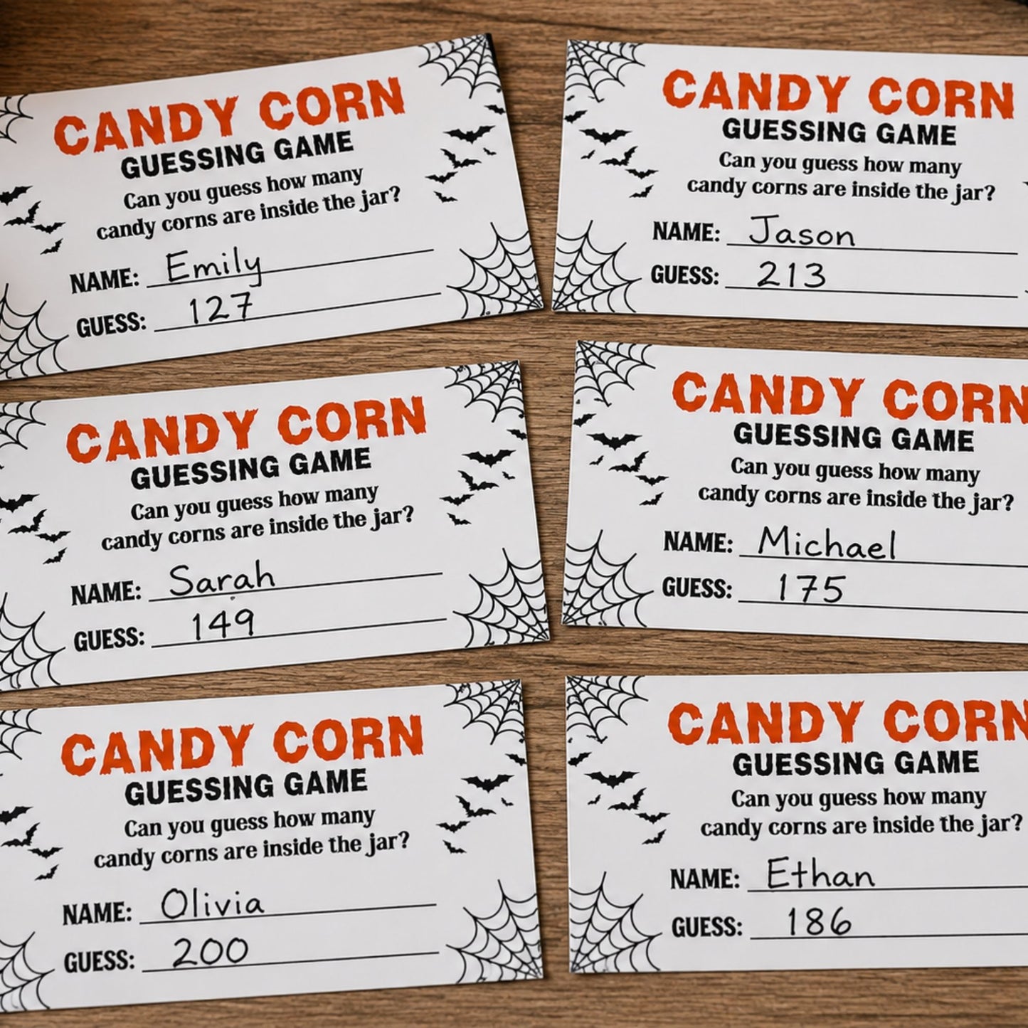 Six Halloween-themed candy corn guessing game cards arranged on a wooden surface. The white cards feature orange text, black spiderweb graphics, bats, and lines for writing names and guesses.