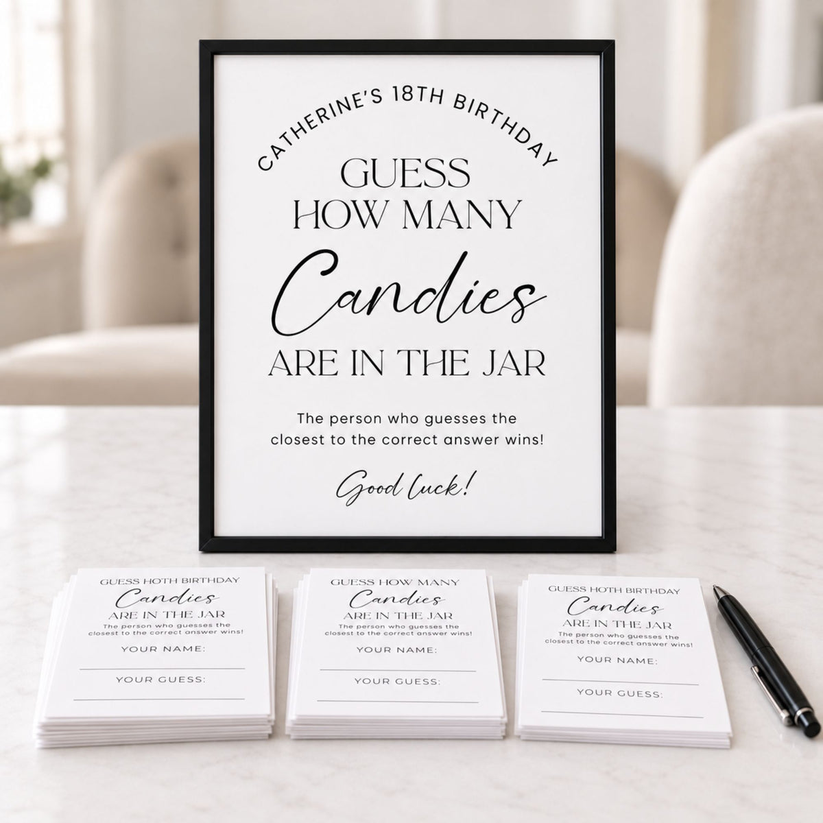 A framed printable party game card reading 'Catherine's 18th Birthday Guess How Many Candies Are in the Jar' is displayed on a light surface, with several matching printed cards and a pen arranged in front. The scene is set in a softly blurred indoor environment with a chair in the background.
