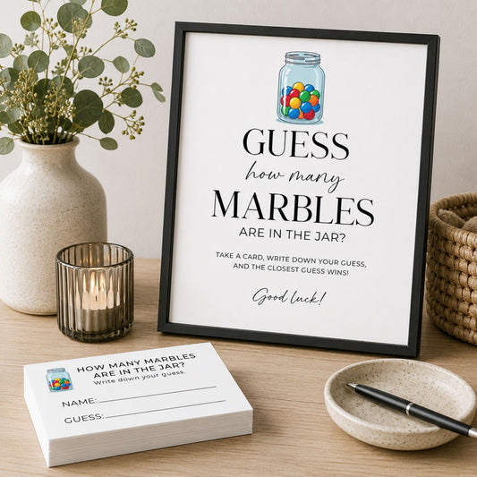 A framed printable party game sign reading 'GUESS how many MARBLES ARE IN THE JAR?' stands on a wooden table beside a card stack, vase with greenery, candle, woven basket, and a small dish with a pen.