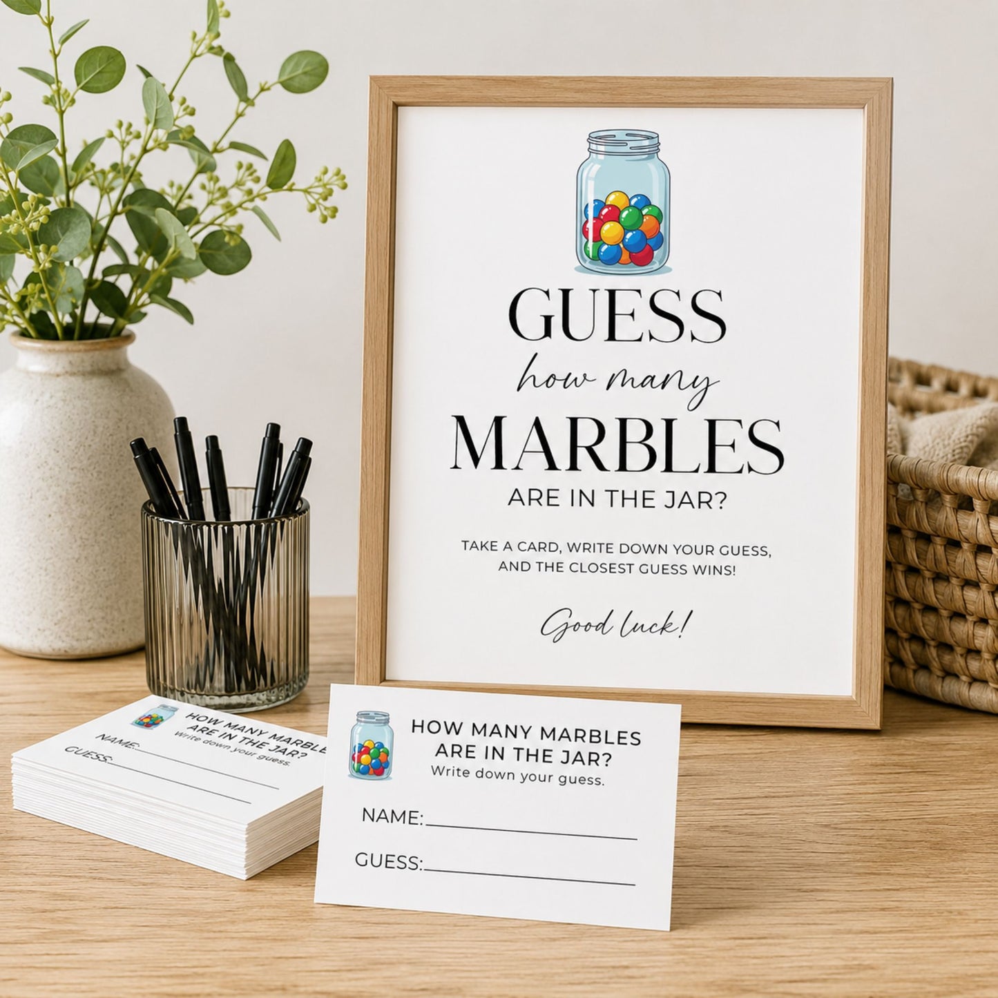 A framed party game print titled 'GUESS how many MARBLES ARE IN THE JAR?' stands on a wooden table, with matching game cards and a small jar of colorful marbles nearby. The scene is styled with a vase of greenery, pens, and a basket in a real indoor setting, so this is Lifestyle rather than Product/Packaging.
