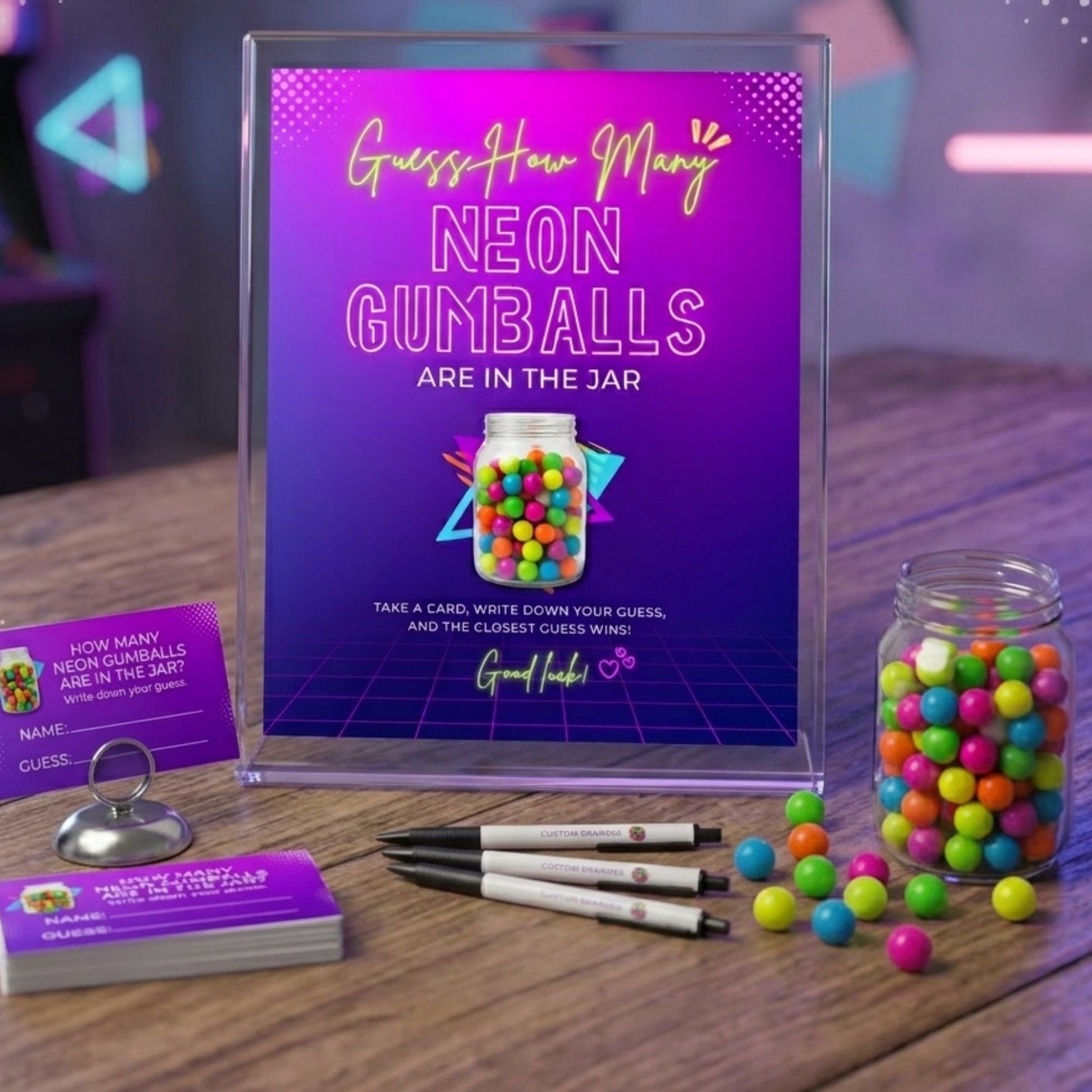 A party game kit for guessing neon gumballs in a jar, featuring a clear acrylic sign with a purple and blue gradient poster, a glass jar filled with colorful gumballs, a stack of purple cards, pens, and a metal ring on a wooden table.