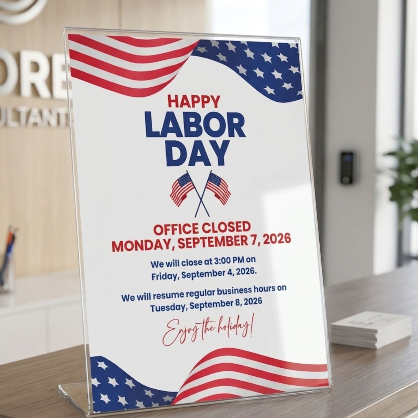 A clear acrylic sign holder displaying a Labor Day office closure notice with American flag graphics, standing on a wooden desk in an office setting.