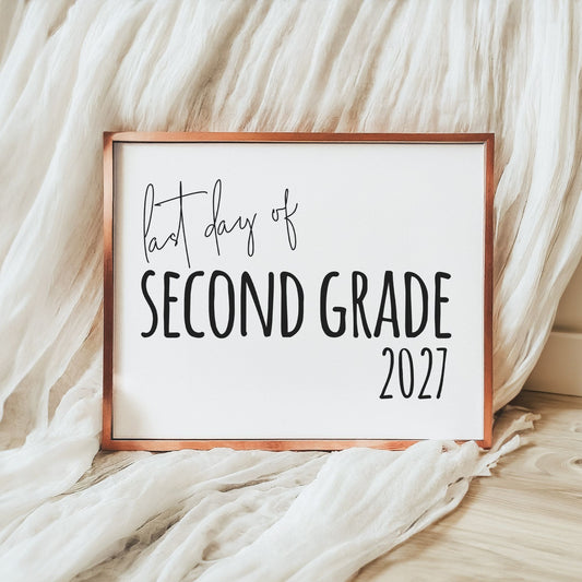 A framed sign reading 'last day of SECOND GRADE 2027' in black script and block lettering on a white background, with a copper-toned frame, styled on a light wood floor with draped white fabric.