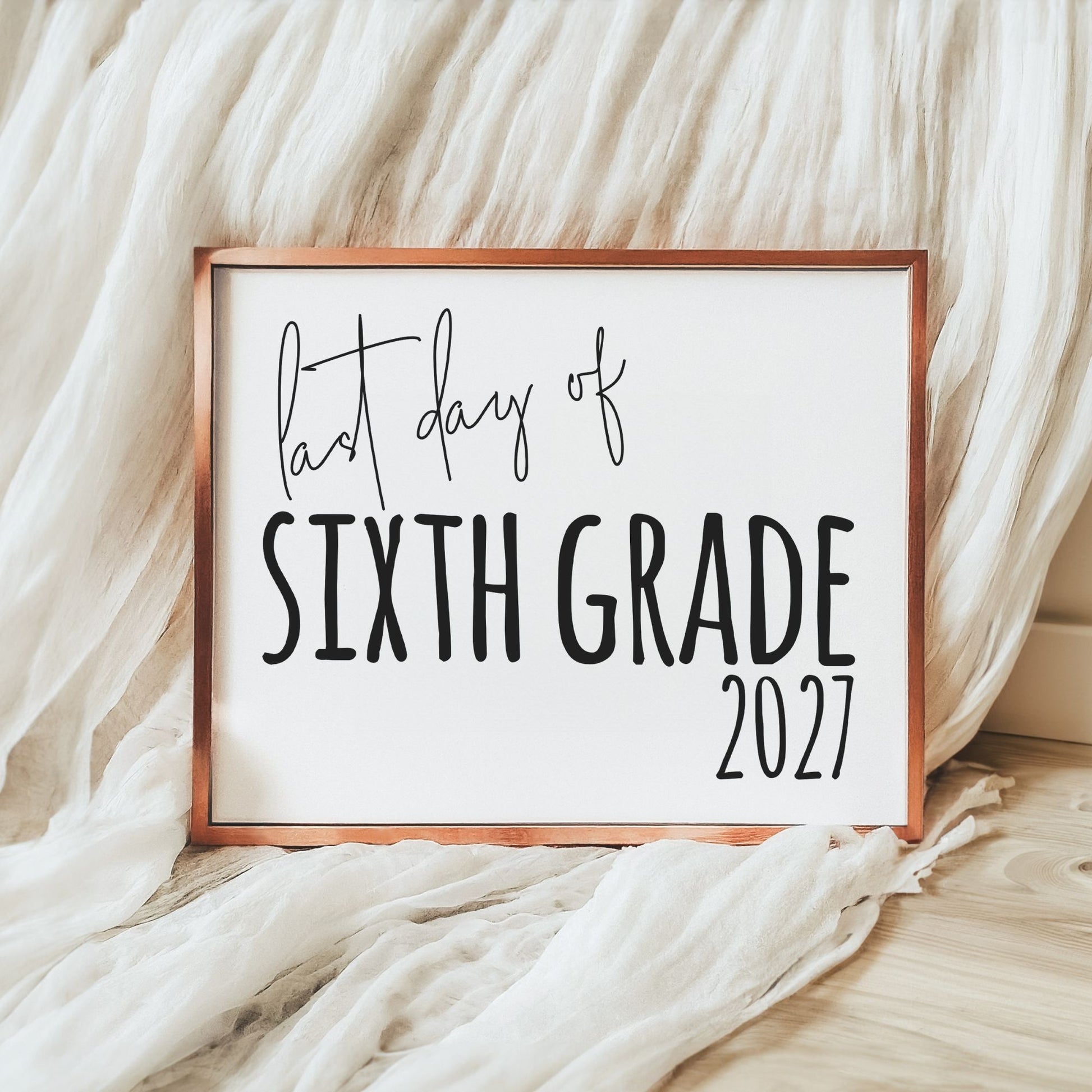 A framed sign reading 'last day of SIXTH GRADE 2027' in black lettering on a white background, with a copper-toned frame, styled on a light wood floor with draped white fabric.