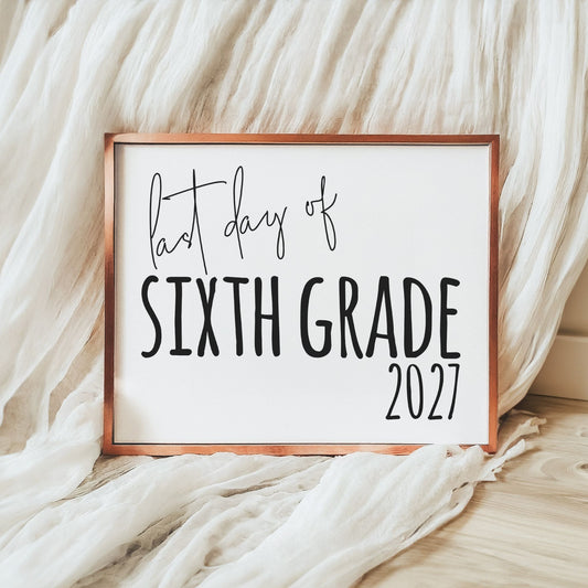 A framed sign reading 'last day of SIXTH GRADE 2027' in black lettering on a white background, with a copper-toned frame, styled on a light wood floor with draped white fabric.