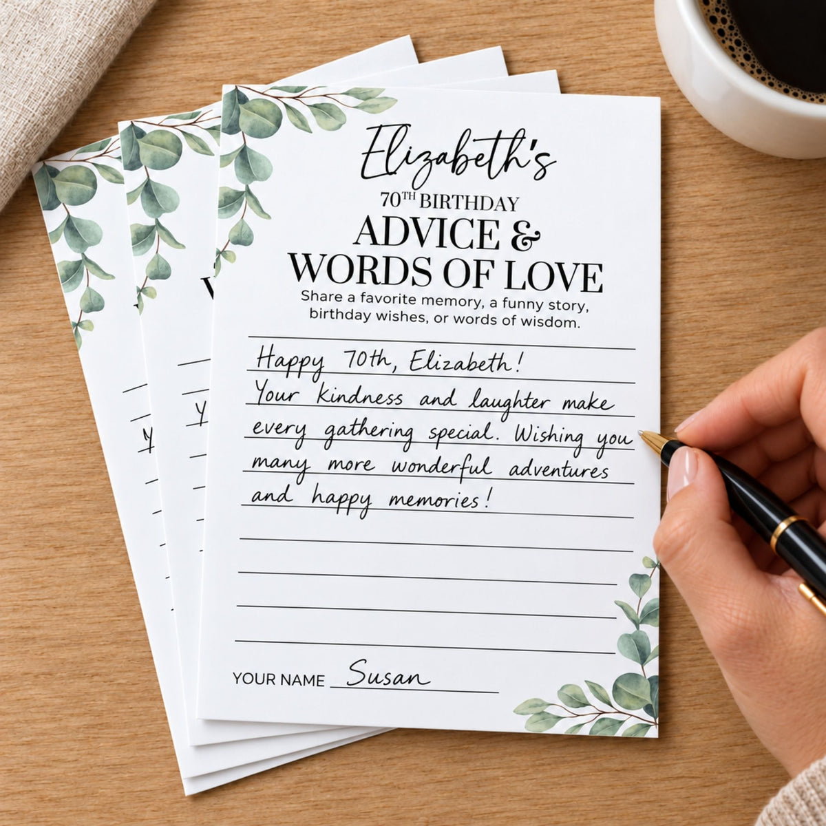 A stack of personalized 70th birthday advice cards with green leaf borders, shown on a wooden table with a hand holding a pen writing on the front card. A coffee cup and fabric are partially visible in the background.