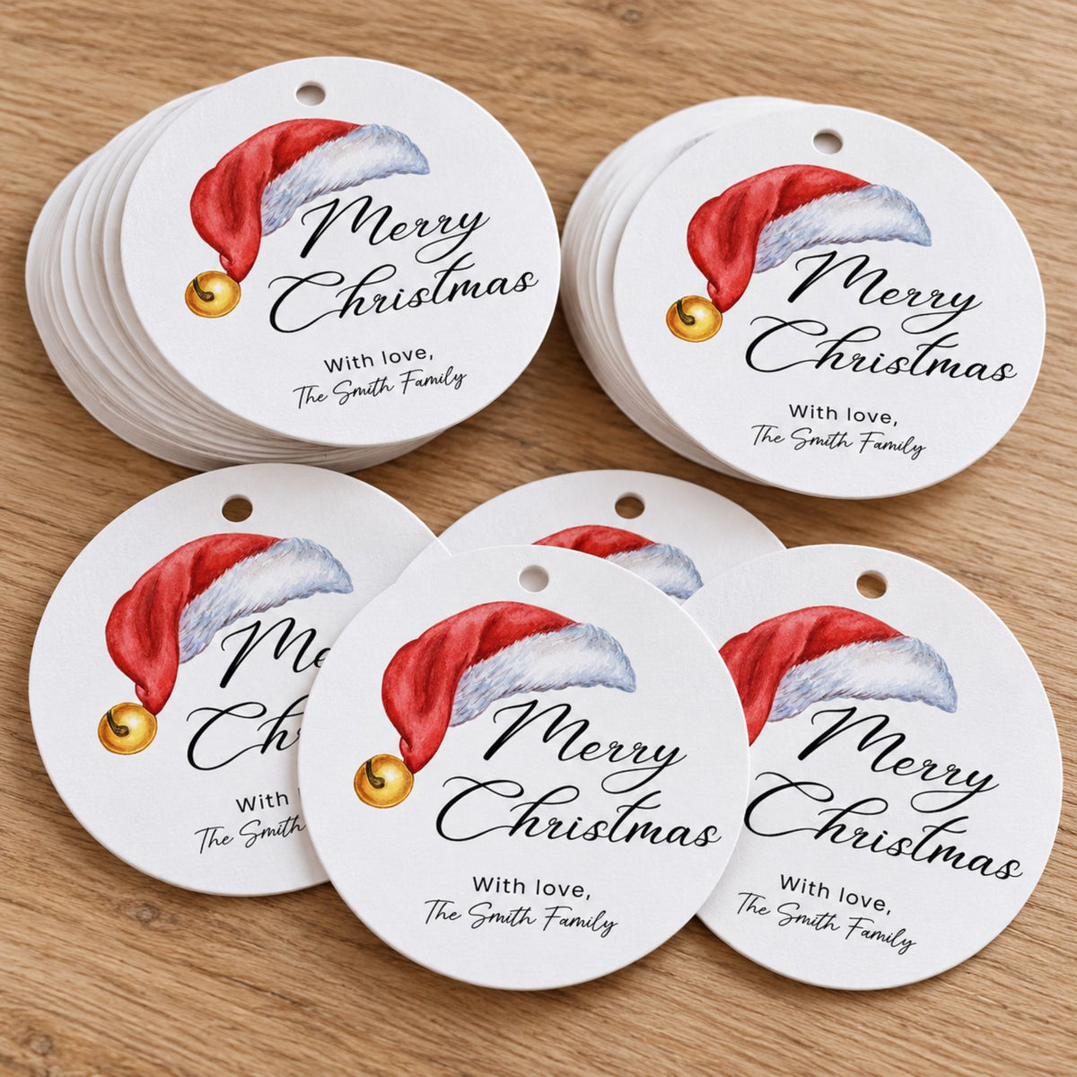 A stack of round white Christmas gift tags with a printed red Santa hat, gold bell, and the text 'Merry Christmas with love, The Smith Family' on a light wood surface.