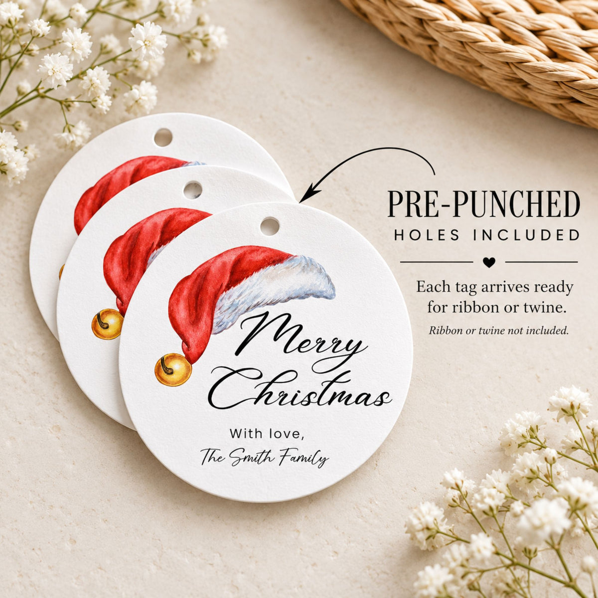 Three round white Christmas gift tags with pre-punched holes, featuring an illustrated red Santa hat and the text 'Merry Christmas' and 'With love, The Smith Family,' arranged on a light beige surface with small white flowers and a woven basket edge.