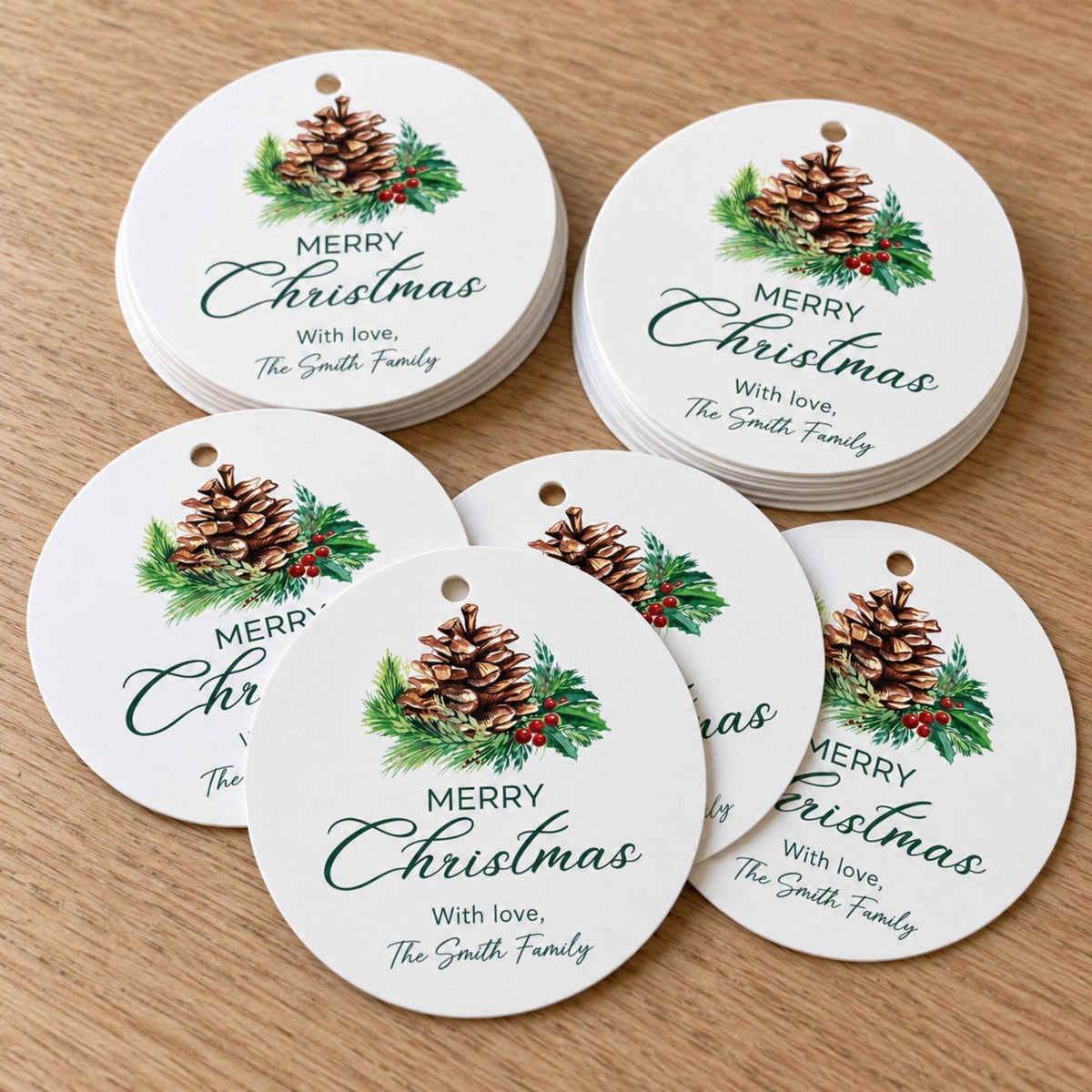 Six round white Christmas ornament tags with a pinecone and green holly design, printed with the text 'MERRY Christmas With love, The Smith Family,' arranged on a light wood surface.