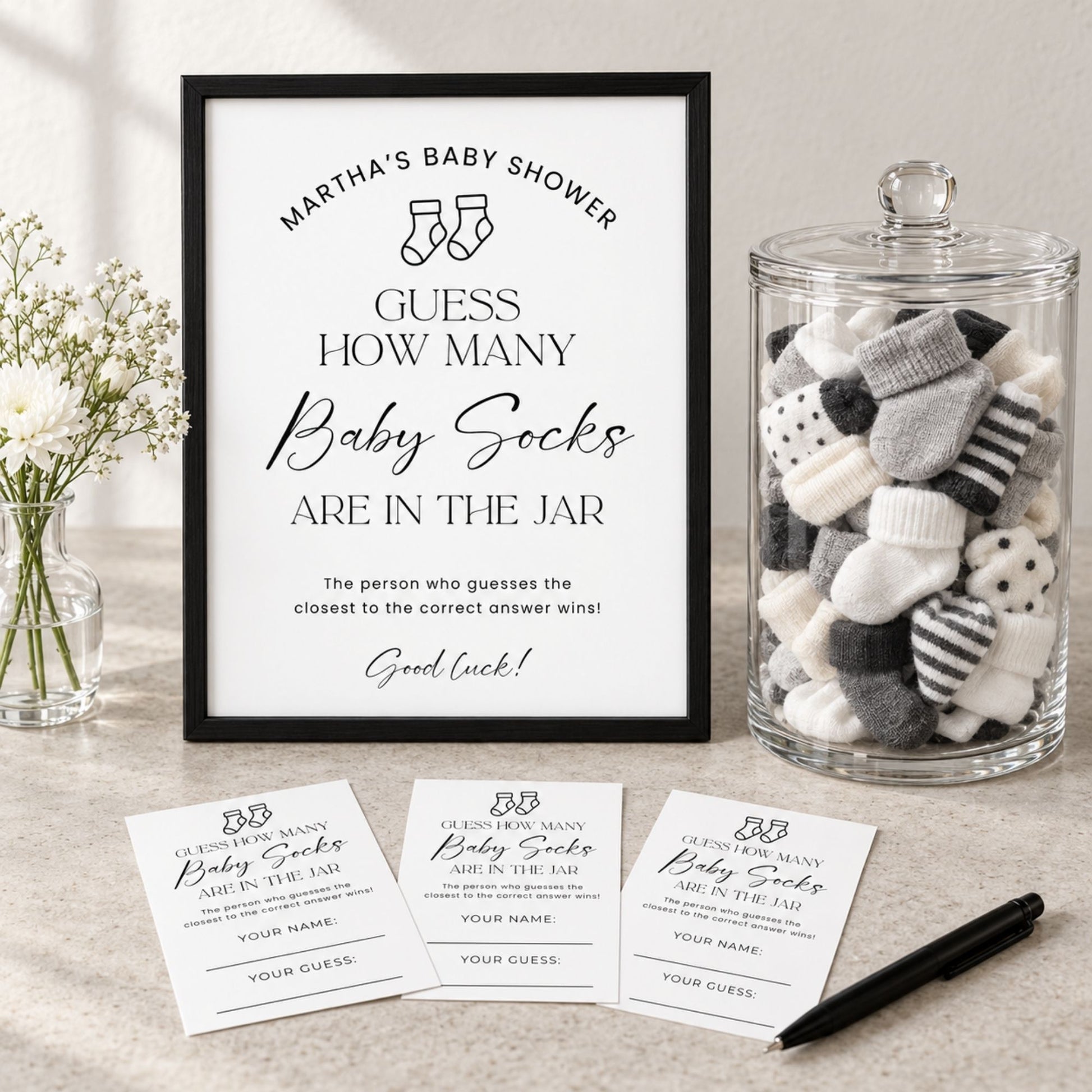 A framed baby shower game sign reading 'Martha's Baby Shower Guess How Many Baby Socks Are in the Jar' is displayed on a tabletop beside a glass jar filled with baby socks, with three matching ticket cards and a pen in front.
