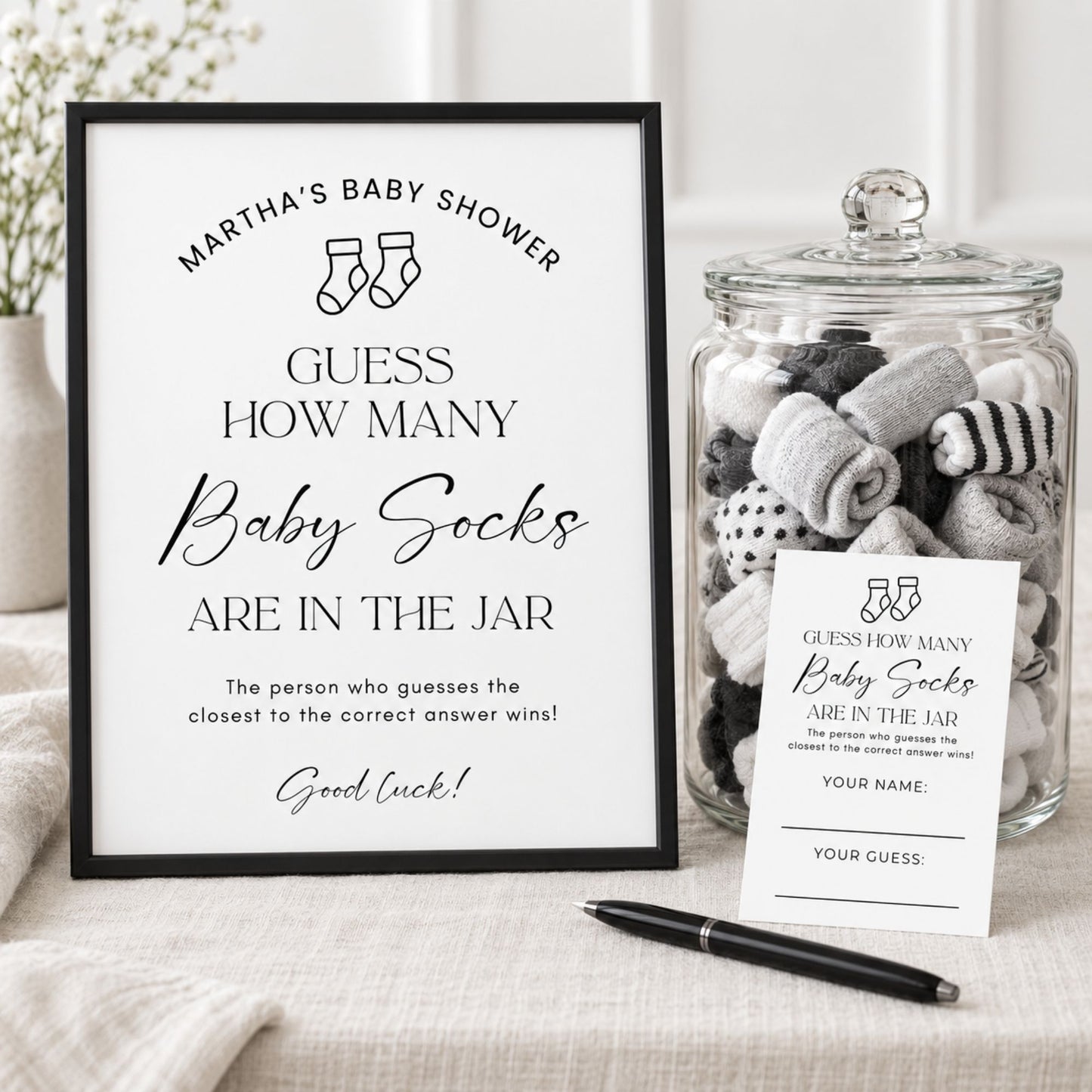 A baby shower game setup featuring a black-framed sign that reads 'Martha's Baby Shower - Guess How Many Baby Socks Are in the Jar,' a clear glass jar filled with rolled baby socks, a small printed card, and a black pen on a light fabric surface.