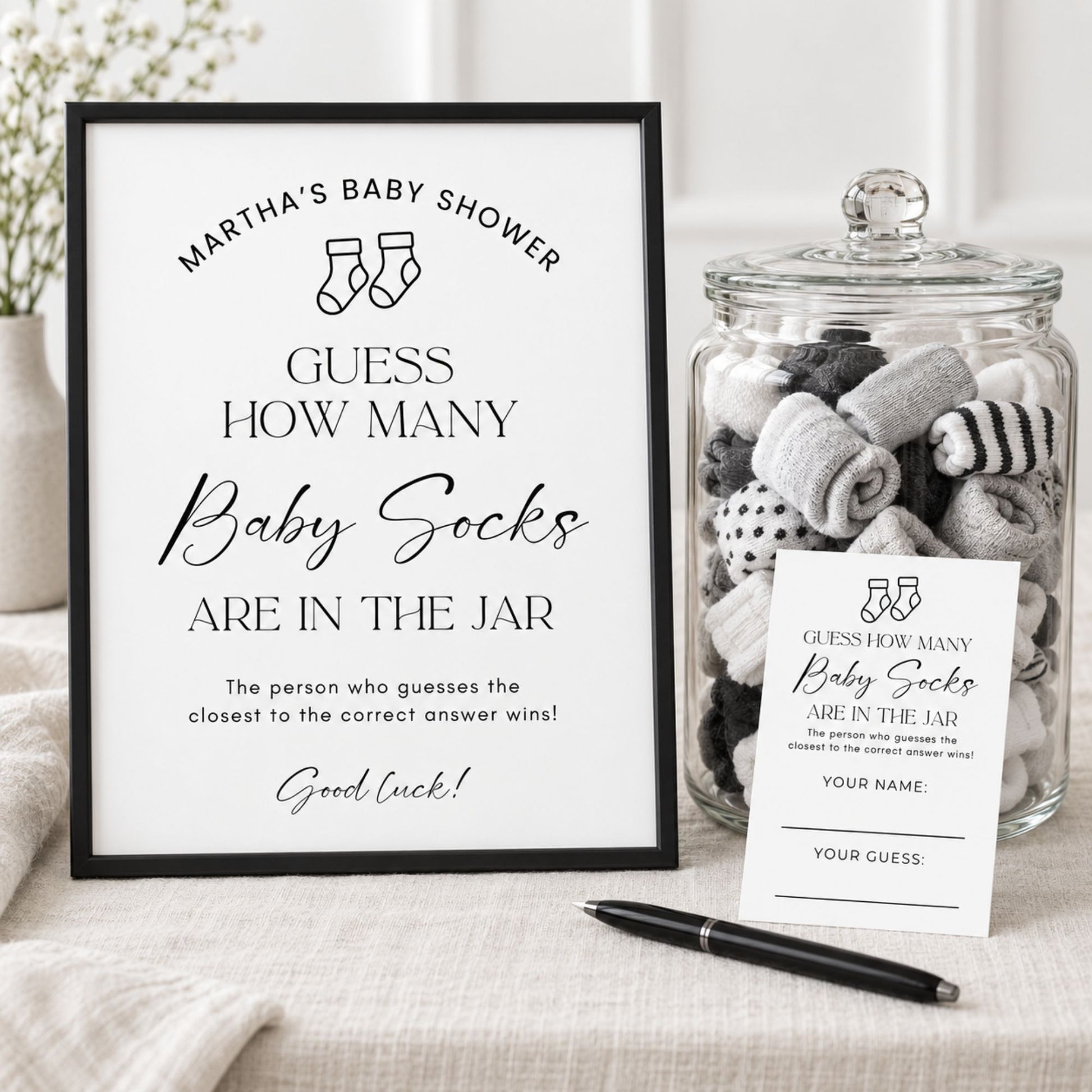 A baby shower game setup featuring a black-framed sign that reads 'Martha's Baby Shower - Guess How Many Baby Socks Are in the Jar,' a clear glass jar filled with rolled baby socks, a small printed card, and a black pen on a light fabric surface.