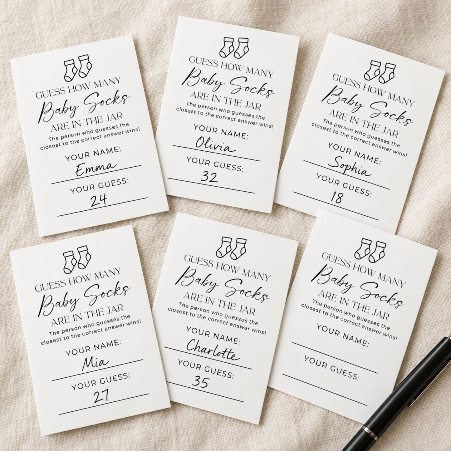 Six white rectangular baby shower game cards with black text and line art, arranged on a light fabric background with a black pen at the bottom right.