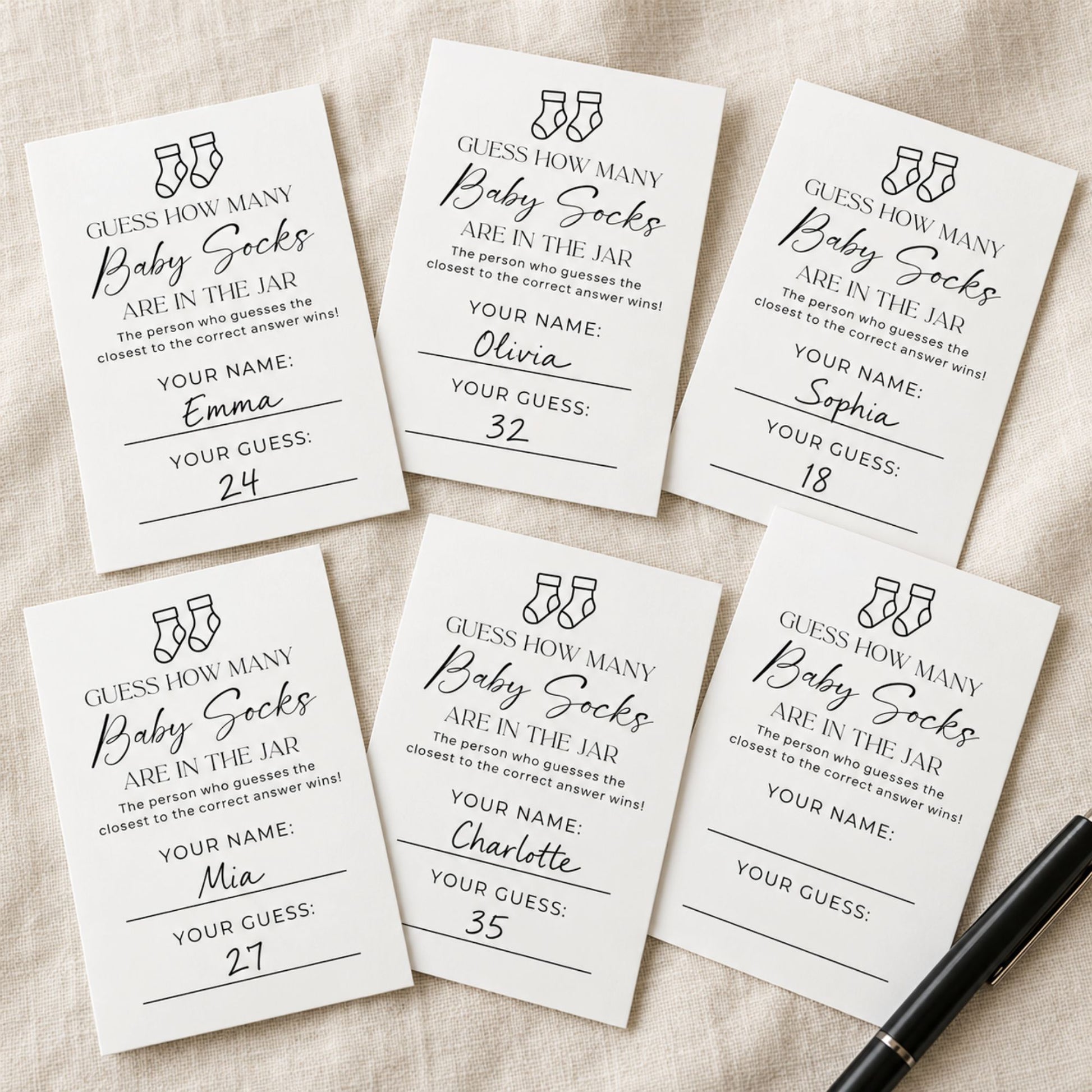 Six white rectangular baby shower game cards with black text and line art, arranged on a light fabric background with a black pen at the bottom right.
