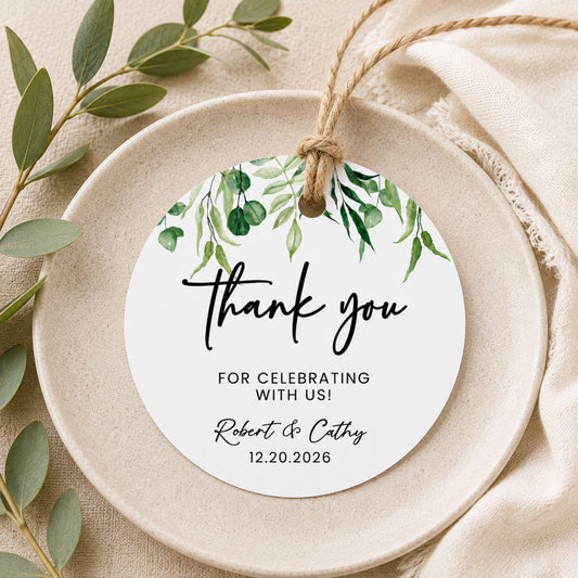 A round white hanging tag with green eucalyptus leaf artwork at the top and the words 'thank you' in black script, styled on a beige ceramic plate with eucalyptus leaves and cream fabric.