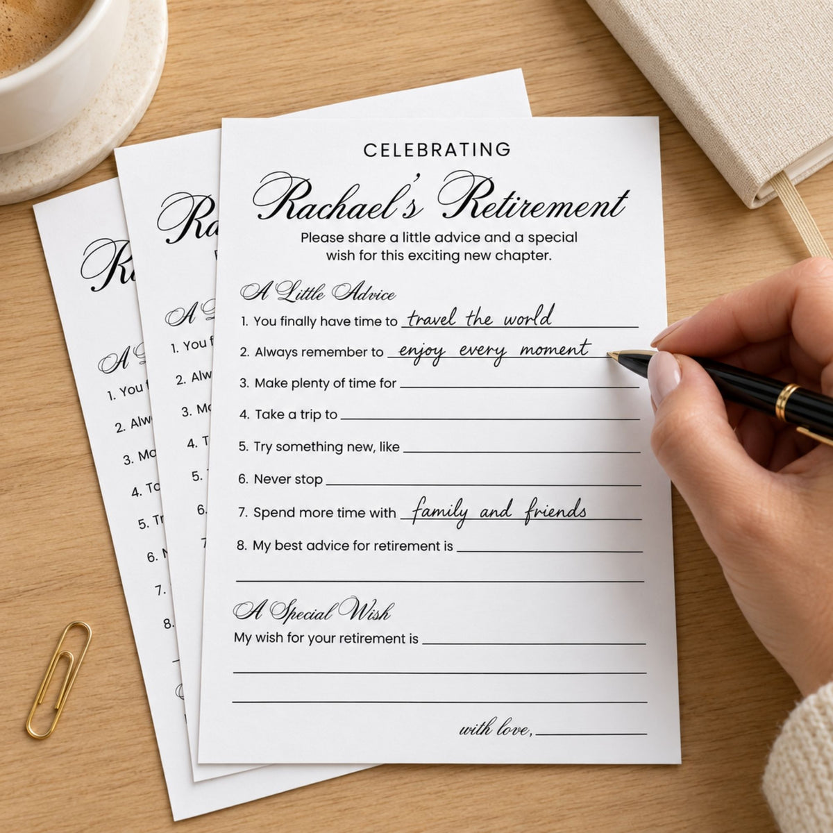 A close-up of a hand writing on a printed retirement reflection card with a black pen, placed on a wooden desk with stacked cards, a coffee cup, and a notebook nearby.