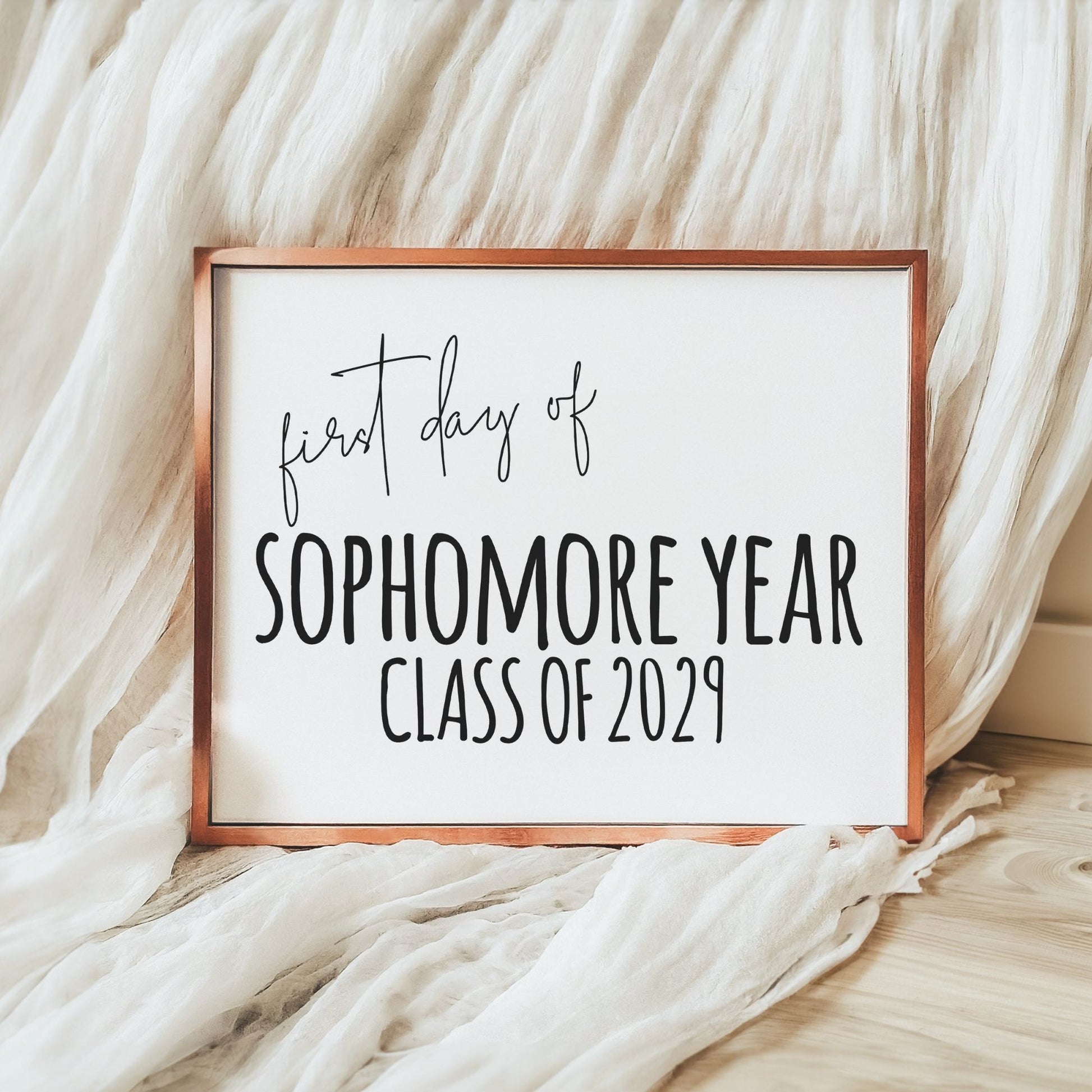 A square framed sign with a copper-toned frame and white background, reading 'first day of SOPHOMORE YEAR CLASS OF 2029' in black script and block lettering, styled on a light wood floor with draped white fabric.