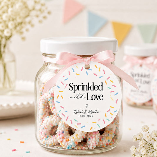 A clear glass jar filled with sugar-dusted pretzels topped with pastel sprinkles, sealed with a white lid and tied with a pink ribbon. A round white tag reading 'Sprinkled with Love' with personalized names and a date hangs from the ribbon.