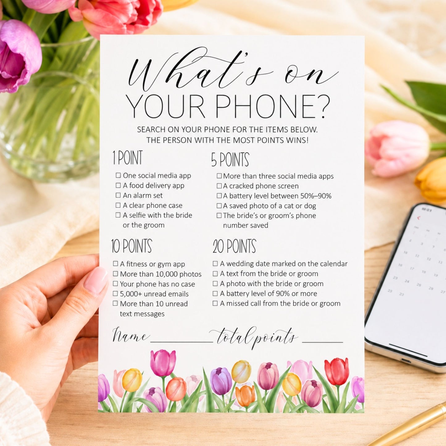 A bridal shower game card titled 'What's on your phone?' held by a hand over a floral table setting with a notebook nearby.